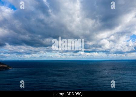 Das riesige blaue Meer erstreckt sich bis zum Horizont unter einem Himmel voller dicker, strukturierter Wolken. Die stimmungsvolle Atmosphäre wird durch das Verschieben von Licht und tiefen Schatten über dem Wasser verstärkt. Stockfoto