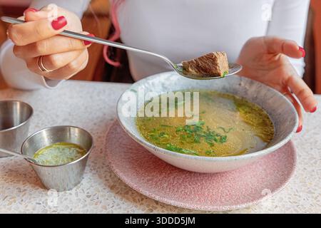 Frau, die dampfende Rinderbrühe schmeckt, einen Löffel zartes Fleisch aus einer warmen Schüssel mit heißer hausgemachter Suppe hebt Stockfoto