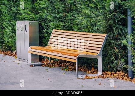 Moderne Parkbank mit einem Recyclingbehälter entlang einer grünen Hecke, mit herabfallenden Herbstblättern auf dem Boden Stockfoto