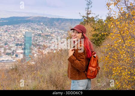Eine Frau, die auf einem malerischen Bergweg unterwegs ist, genießt einen Panoramablick auf die Stadt und das lebhafte Herbstlaub während ihrer Alleinreise Stockfoto