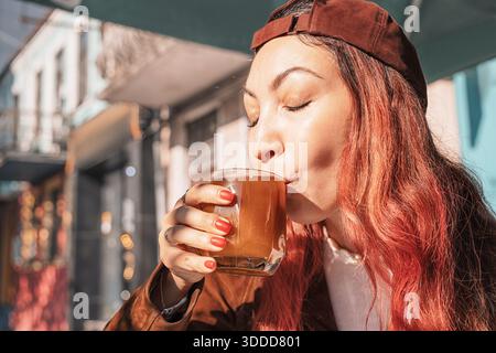 Junge Frau mit roten Haaren, die die Augen schließen, während sie ein heißes Getränk in einem Becher genießt, in einem Café im Freien sitzt und sich entspannt Stockfoto