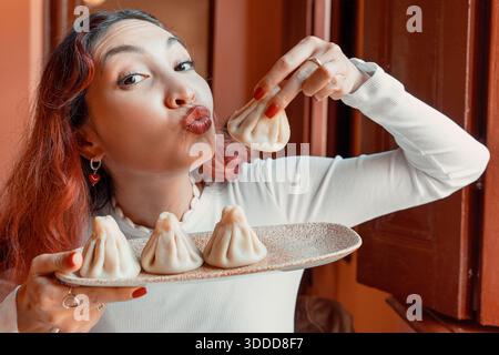 Eine junge Frau, die einen Teller traditioneller georgischer Chinkali hält, die Lippen spielerisch zusammendrückt und einen Knödel hebt Stockfoto