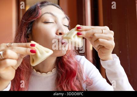 Eine junge Frau, die einen Teller traditioneller georgischer Chinkali hält, die Lippen spielerisch zusammendrückt und einen Knödel hebt Stockfoto