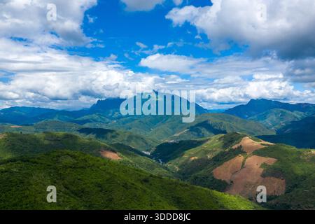 Weitläufige grüne Hügel und bewaldete Hänge erstrecken sich in Richtung einer dramatischen Bergkette unter einem leuchtend blauen Himmel mit verstreuten Wolken im Norden Laos. Die Landschaft bietet Flächen mit bewirtschaftetem Land, sanftem Gelände und ein Gefühl von ungeheurer natürlicher Offenheit. Stockfoto