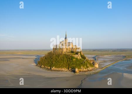 Blick aus der Vogelperspektive auf die Abtei Mont-Saint-Michel auf der felsigen Insel, umgeben von weiten Wattflächen und sanfter Morgensonne unter einem klaren blauen Himmel. Die Szene zeigt die ikonische mittelalterliche Architektur und die ruhige, offene Landschaft. Stockfoto