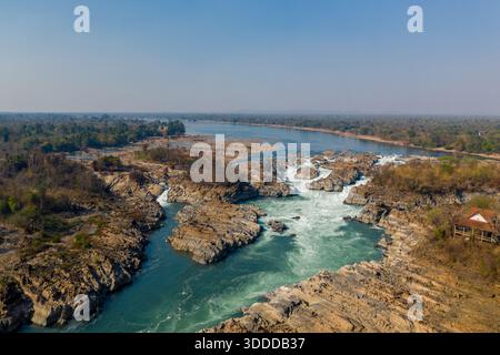 Ausgedehnte Luftszene der Khone Phapheng Wasserfälle am Mekong River im Süden von Laos. Turbulentes Weißwasser stürzt über zerklüftete Felsen, umgeben von trockenem Wald und einer breiten Flusslandschaft unter einem klaren blauen Himmel. Stockfoto
