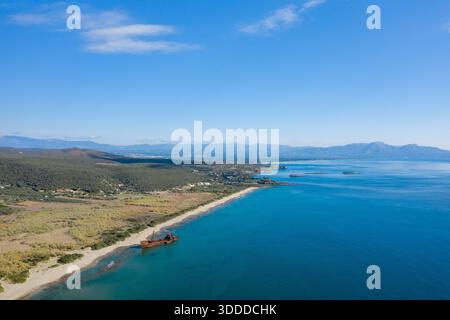 Aus der Vogelperspektive auf das Schiffswrack von Dimitrios, das an einem langen Sandstrand mit türkisfarbenem Meer und üppigen Hügeln in der Nähe von Gythio, Griechenland, ruht. Helles Sonnenlicht und ein großer blauer Himmel schaffen eine friedliche, weitläufige Küstenlandschaft. Stockfoto