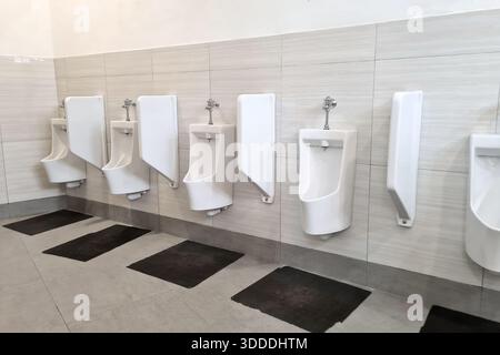 Saubere, moderne öffentliche Toiletten mit einer Reihe von an der Wand montierten Urinalen mit minimalistischem Design, gefliesten Wänden und hygienischem Layout, die für sanitäre Einrichtungen stehen Stockfoto