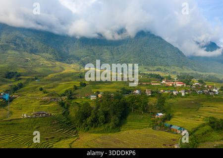Ein kleines Dorf eingebettet zwischen lebendigen Reisterrassen-Reisfeldern am Fuße der üppig grünen Berge in Sapa, Vietnam. Niedrige Wolken ziehen über die Gipfel und verleihen der ländlichen Landschaft eine dramatische und ruhige Atmosphäre. Stockfoto