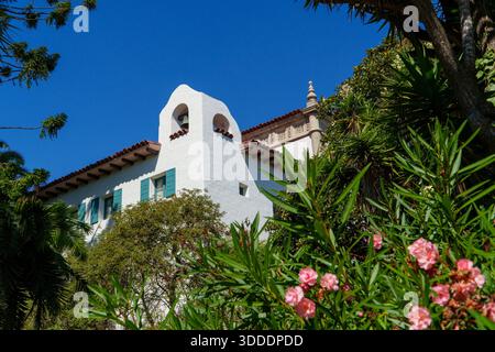 Ein weißes architektonisches Gebäude mit Glockenturm, rotem Ziegeldach und blauen Fensterläden ist von lebhaftem tropischem Grün und rosa Oleanderblüten eingerahmt Stockfoto