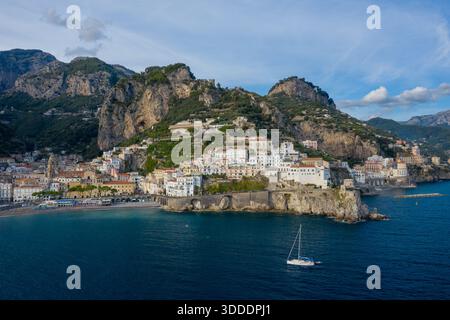 Ein weißes Segelboot schwimmt auf tiefblauem Mittelmeer direkt vor der Küste der sonnendurchfluteten Stadt Amalfi, Italien. Die Szene bietet pastellfarbene Gebäude, dramatische felsige Klippen und üppige grüne Terrassen unter weichem Himmel. Stockfoto