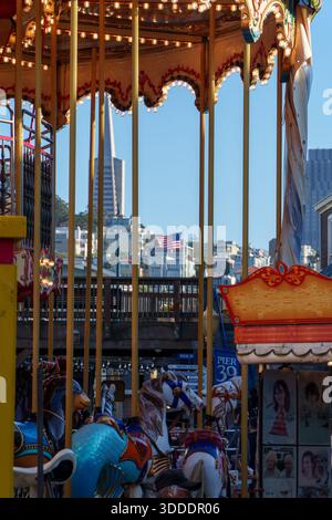 Am Pier 39 steht ein farbenfrohes Karussell mit goldenen Stangen und leuchtenden Lichtern, das die Skyline von San Francisco und die amerikanische Flagge umrahmt. Stockfoto