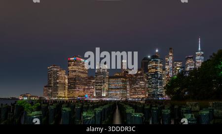Die beleuchtete Skyline von Lower Manhattan mit markanten Wolkenkratzern und dem One World Trade Center erhebt sich nachts über dunklen Holzpfeilern. Stockfoto