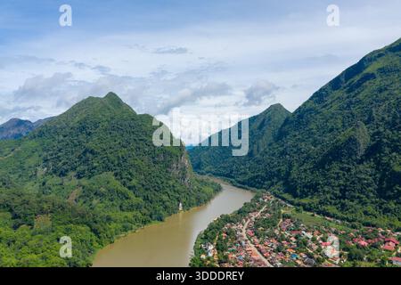 Eine atemberaubende Luftperspektive fängt den Fluss Nam ou ein, der sich durch die üppig grünen Kalksteinberge schlängelt und das Dorf Nong Khiaw in Laos. Der dichte tropische Wald und die gewachsenen Dächer sorgen für eine lebhafte Atmosphäre unter einem hellen Sommerhimmel. Stockfoto