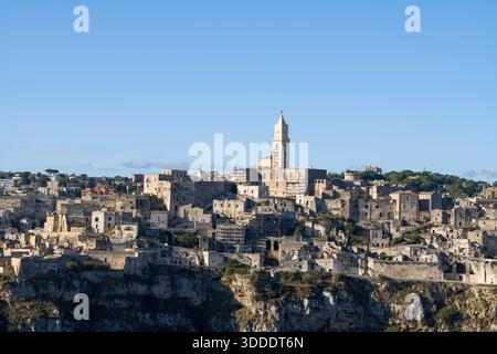 Historische Steinbauten von Matera erheben sich über zerklüftete Klippen, wobei der markante Turm der Kathedrale vor einem klaren blauen Himmel steht und das Sonnenlicht die antike Architektur hervorhebt. Stockfoto