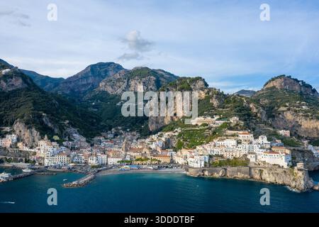 Ein weitläufiger Blick aus der Vogelperspektive fängt die Stadt Amalfi, Italien, mit weiß getünchten Gebäuden entlang der dramatischen Küste und umgeben von steilen, felsigen Bergen ein. Die Szene bietet klares mediterranes Wasser, Terrassengrün und eine ruhige Atmosphäre unter einem sanften blauen Himmel. Stockfoto