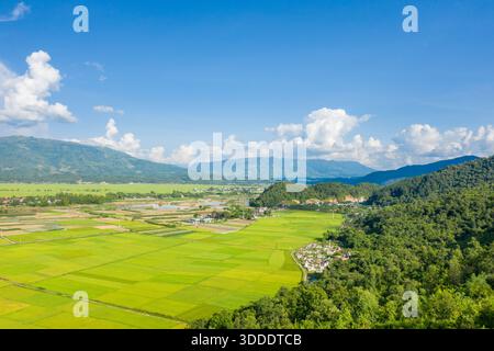 Ausgedehnte Reisfelder erstrecken sich über ein üppiges Tal, das von bewaldeten Hügeln und fernen blauen Bergen in der Nähe von Dien Bien Phu umgeben ist. Helles Sonnenlicht und verstreute Cumulus-Wolken schaffen eine frische, lebendige ländliche Landschaft. Stockfoto