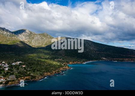 Eine zerklüftete Küste mit tiefblauem Wasser kurvt unter steilen, felsigen Bergen, die von Grün bedeckt sind. Sonnenlicht und Schatten spielen in der Landschaft unter einem Himmel voller strukturierter Wolken. Stockfoto