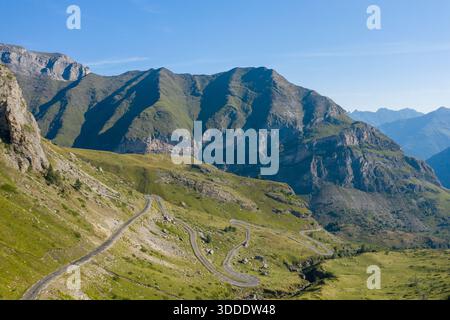 Kurvenreiche Bergstraße schlängelt sich über grasbewachsene Hänge und felsiges Gelände in den Pyrenäen bei Gavarnie Gedre. Helles Tageslicht unterstreicht die dramatischen Konturen und zerklüfteten Klippen unter einem klaren blauen Himmel. Stockfoto