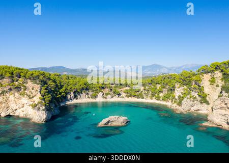 Weite Panoramaaufnahme einer abgeschiedenen Bucht mit kristallklarem türkisfarbenem Wasser, umgeben von zerklüfteten Klippen und dichtem Kiefernwald am Alonaki Fanariou Strand in Griechenland. Helles Sonnenlicht und ein wolkenloser Himmel unterstreichen die lebendige mediterrane Landschaft. Stockfoto