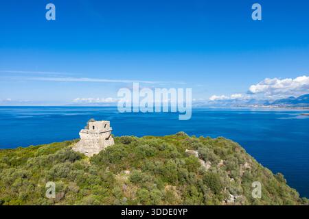 Der alte Wachturm aus Stein erhebt sich über der dichten grünen Vegetation auf der Isola di Cirella, umgeben von tiefblauen mediterranen Gewässern und fernen Bergen unter klarem Himmel. Helles Sonnenlicht verstärkt die rauen Strukturen und die ruhige Küstenatmosphäre. Stockfoto
