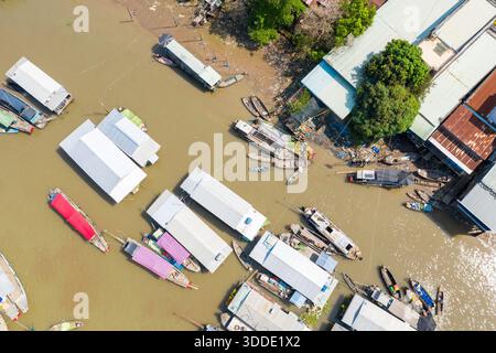 Aus der Sicht von oben zeigt sich ein geometrisches Muster von Marktbooten mit farbenfrohen Metalldächern, die entlang des Flusses am schwimmenden Markt Cai Rang in Can Tho, Mekong Delta, Vietnam, vertäut sind. Das Bild hebt den Kontrast zwischen den geclusterten Schiffen, den Strukturen am Fluss und den üppigen grünen Bäumen bei hellem Tageslicht hervor. Stockfoto