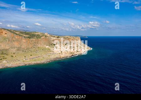 Blick aus der Vogelperspektive auf das historische Dorf Monemvasia, eingebettet unter steilen orangen Klippen am leuchtend blauen Mittelmeer. Die Szene bietet zerklüftete Felsstrukturen, klares Wasser und einen hellen Himmel mit verstreuten Wolken. Stockfoto