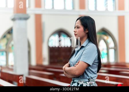 Seitenansicht einer religiösen Frau, die mit gefalteten Händen betet, in einer Kirche mit bogenförmigen Fenstern im Hintergrund. Stockfoto