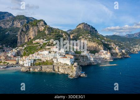 Weißgetünchte Gebäude in Amalfi, Italien, stürzen sich über steile Klippen hinunter und überblicken das tiefblaue Mittelmeer. Die dramatische Küste wird von üppigen grünen Hügeln und zerklüfteten Bergen unter einem klaren Himmel umgeben. Stockfoto
