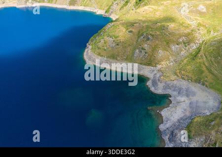 Bild über dem Kopf des lebendigen blauen Wassers des Lac Bleu, das auf felsige und grasbewachsene Hänge in den Pyrenäen trifft. Helles Sonnenlicht verstärkt die kontrastierenden Strukturen und Farben entlang des Seeufers. Stockfoto