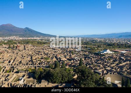 Weite Aussicht aus der Vogelperspektive auf die antiken Steinruinen von Pompeji mit dem Vesuv, der sich im Hintergrund unter einem leuchtend blauen Himmel erhebt. Klares Sonnenlicht hebt archäologische Strukturen, verstreutes Grün und die dramatische vulkanische Landschaft Süditaliens hervor. Stockfoto