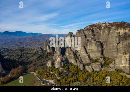 Das Kloster Varlaam thront auf einer dramatischen Sandsteinsäule, umgeben von Herbstwäldern und zerklüfteten Bergen in Meteora, Griechenland. Die Szene zeigt weitläufige Felsformationen unter einem leuchtend blauen Himmel, die ein Gefühl von Pracht und Isolation schaffen. Stockfoto