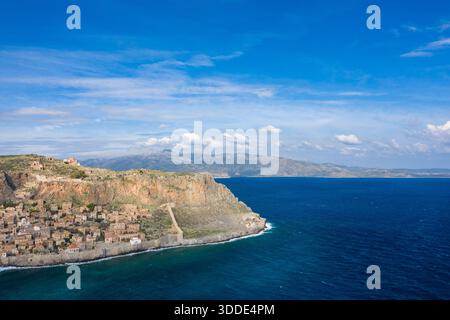 Weite Aussicht aus der Vogelperspektive auf Monemvasias historisches Steindorf unter zerklüfteten Klippen mit Blick auf das lebendig blaue Mittelmeer unter einem hellen Himmel mit verstreuten Wolken. Stockfoto
