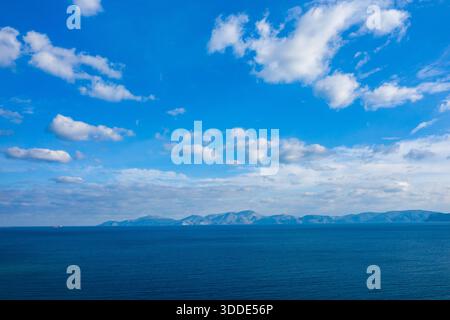 Weite Ausdehnung des tiefblauen Meeres erstreckt sich in Richtung einer fernen Bergkette unter einem Himmel voller verstreuter weißer Wolken. Die ruhige Szene zeichnet sich durch subtile Verläufe und eine friedliche, offene Atmosphäre aus. Stockfoto