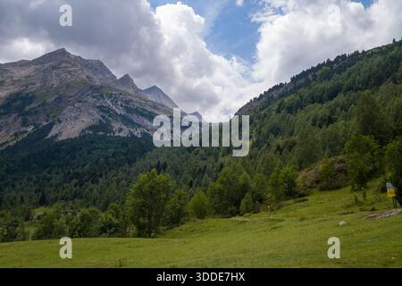 Ein markanter felsiger Bergkamm erhebt sich über einem üppig grünen Tal mit dichtem Wald unter einem teilweise bewölkten Himmel in den Pyrenäen. Die Landschaft zeichnet sich durch mehrschichtige Strukturen, lebhaftes Grün und dynamisches Sommerlicht aus. Stockfoto