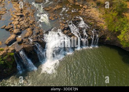 Die atemberaubende Perspektive des Wasserfalls Tad Lo, während das Wasser über zerklüftete Felsen in ein breites Becken im Süden von Laos stürzt. Sonnendurchflutete Texturen heben den Kontrast zwischen fließendem Wasser, freiliegendem Stein und umliegendem Grün hervor. Stockfoto