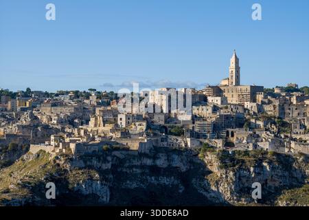 Historische Steinbauten und der Turm der Kathedrale von Matera, Italien, erheben sich über dramatischen Klippen, die von warmem Tageslicht unter einem klaren blauen Himmel beleuchtet werden. Stockfoto