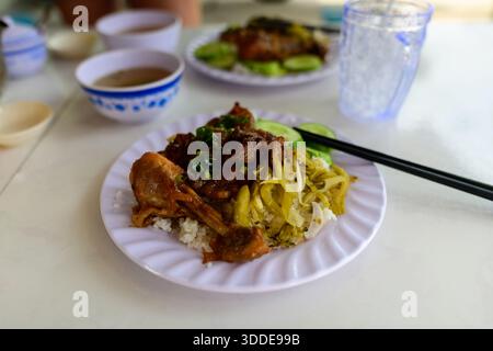 Eine Nahaufnahme eines traditionellen vietnamesischen Reisgerichts mit geschmortem Fleisch, eingelegtem Senfgrün und geschnittener Gurke auf einem weißen Teller. Die Mahlzeit wird mit Essstäbchen serviert und von Schüsseln Suppe und Eiswasser auf einem einfachen Tisch begleitet. Stockfoto