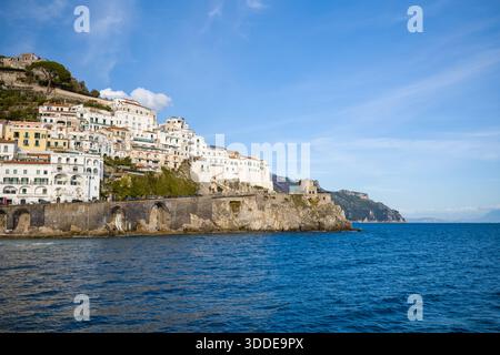 In Amalfi, Italien, befinden sich sonnendurchflutete weiße und pastellfarbene Gebäude, die entlang steiler Klippen mit Blick auf das tiefblaue Mittelmeer gestapelt sind. Die dramatische Küste bietet historische Steinbögen und einen mittelalterlichen Wachturm unter klarem Himmel. Stockfoto