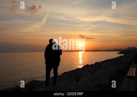 Die Silhouette eines Mannes, der an einer felsigen Küste steht und den Sonnenuntergang über einem ruhigen Meer beobachtet Stockfoto