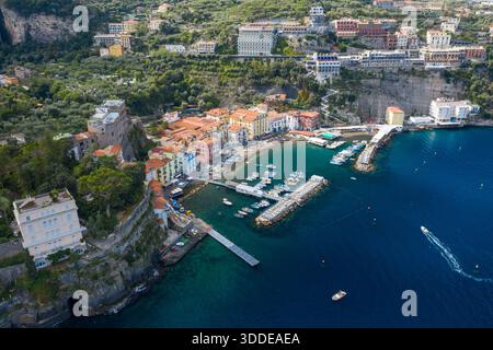 Hochwinkelperspektive von Sorrent, Italien, zeigt einen lebendigen Yachthafen mit Booten, die entlang der Uferpromenade angedockt sind, und pastellfarbene Gebäude, die sich unter dramatischen Klippen gruppieren. Die Szene bietet üppiges Grün, klares blaues Wasser und eine lebhafte mediterrane Küstenlandschaft. Stockfoto