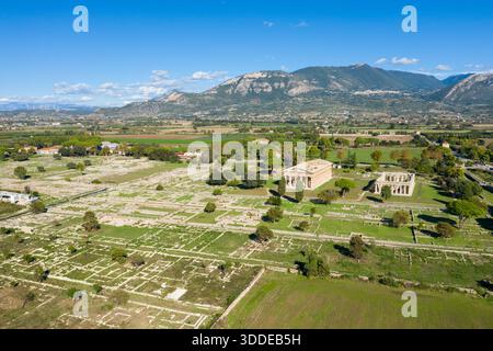 Weite Luftperspektive der antiken archäologischen Stätte Paestum, Italien, mit weitläufigen Steinruinen und zwei gut erhaltenen griechischen Tempeln inmitten grüner Felder. Die Landschaft ist von fernen Bergen und einem leuchtend blauen mediterranen Himmel mit verstreuten Wolken umgeben. Stockfoto