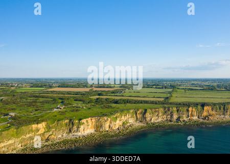 Luftlandschaft mit zerklüfteten goldenen Klippen über einer felsigen Küste, mit üppigen grünen Feldern und Ackerland, die sich bis in die Ferne unter dem klaren blauen Himmel in Pointe du hoc erstrecken. Stockfoto
