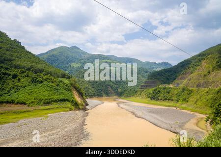 Ein schlammiger Fluss fließt durch ein weites Tal, das von üppigen grünen Bergen im Norden Vietnams flankiert wird. Helles Tageslicht und verstreute Wolken unterstreichen die natürlichen Strukturen der Landschaft. Stockfoto