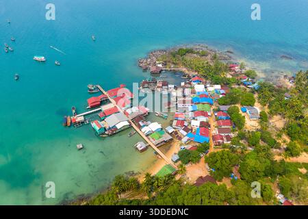 Aus der Vogelperspektive erschließt sich ein lebendiges Fischerdorf mit bunten Dächern, Holzstegen und Booten, die in türkisfarbenem Wasser am Coconut Beach auf Koh Rong Island in Kambodscha verankert sind. Üppige tropische Vegetation und felsige Küste umrahmen diese lebhafte Küstensiedlung. Stockfoto
