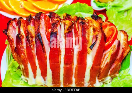 Aus nächster Nähe sehen Sie ein herzhaftes Schweinebauchgericht, das fachmännisch geröstet und mit knusprigen Speckstreifen und frischen Tomatenscheiben garniert wird und auf einem Bett mit knusprigen Tomaten serviert wird Stockfoto