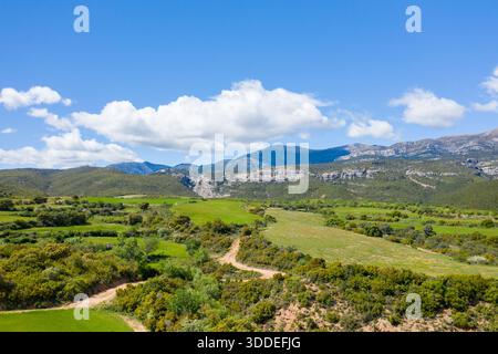 Ein kurviger Schotterweg schlängelt sich durch üppige grüne Felder und niedrige Sträucher und führt zu entfernten felsigen Bergen unter einem leuchtend blauen Himmel in der Nähe von Bierge, Spanien. Geschwollene weiße Wolken werfen weiche Schatten über die lebendige Frühlingslandschaft. Stockfoto