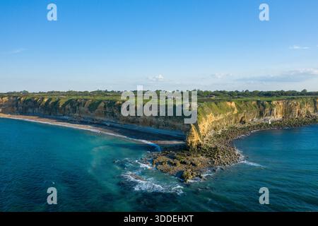 Weite Aussicht aus der Vogelperspektive auf zerklüftete goldene Klippen, die sich über klarem türkisfarbenem Wasser in Pointe du hoc in der Normandie erheben, mit Sonnenlicht, das scharfe Schatten wirft und eine ruhige Küstenatmosphäre bietet. Stockfoto