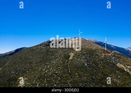Drei Windräder stehen auf einem zerklüfteten, grünen Hügel unter einem klaren blauen Himmel in der Nähe von Thisbe, Griechenland. Die Landschaft zeichnet sich durch spärliche Vegetation, erdige Texturen und gewundene, unbefestigte Straßen bei hellem Tageslicht aus. Stockfoto