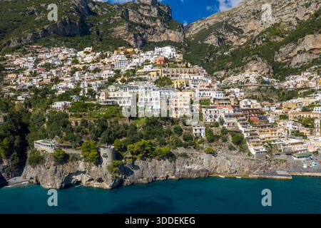 Farbenfrohe mediterrane Gebäude stürzen sich auf den steilen Hügel von Positano, Italien, mit Blick auf türkisfarbenes Wasser und zerklüftete Klippen. Üppiges Grün und ein historischer Wachturm aus Stein unterstreichen die dramatische Küstenlandschaft unter einem klaren blauen Himmel. Stockfoto